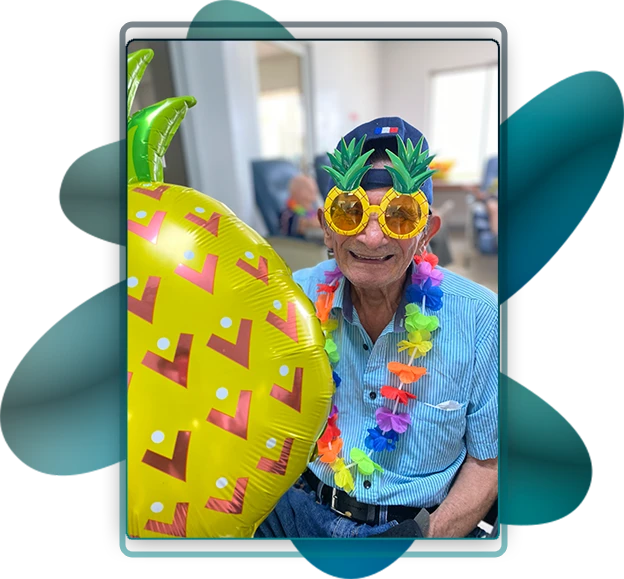 Fotografía de persona adulta mayor, disfrazado con temática de fruta como la piña, el adulto mayor tiene lentes decorados, collar y globo de la piña